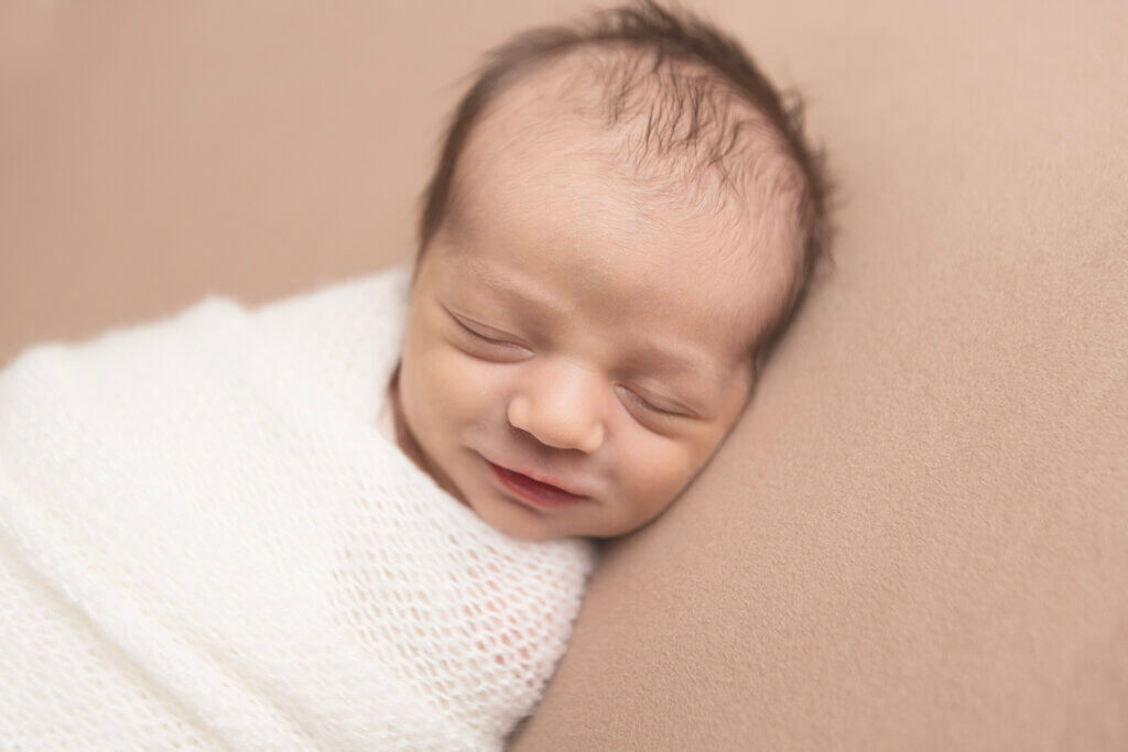 Newborn baby boy peacefully sleeping, wrapped in a soft cream blanket on a neutral backdrop during a studio newborn session in Pawtucket, Rhode Island.