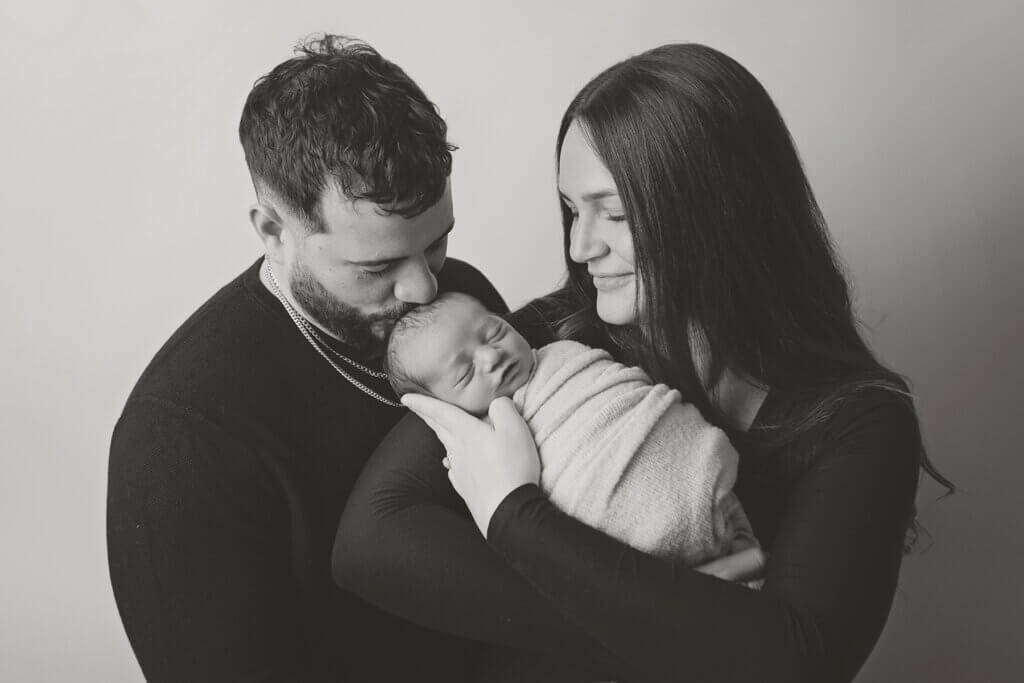 Mom and dad gently kissing their newborn baby’s head during a family newborn photography session in Pawtucket, Rhode Island.