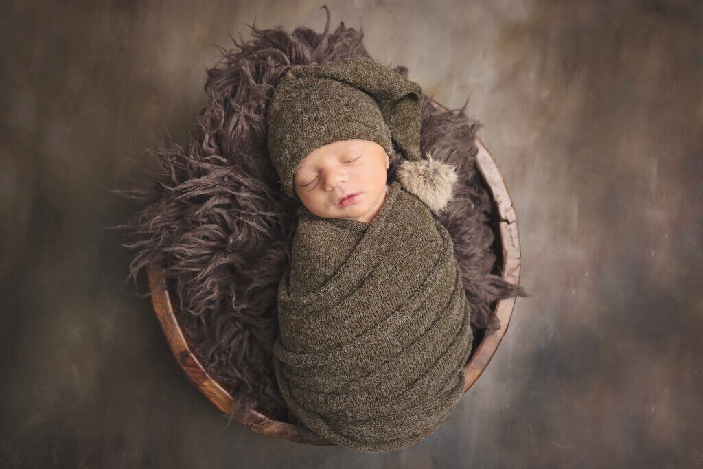 Newborn baby boy posed in a bowl with a green Santa hat and swaddle during a Christmas newborn photography session